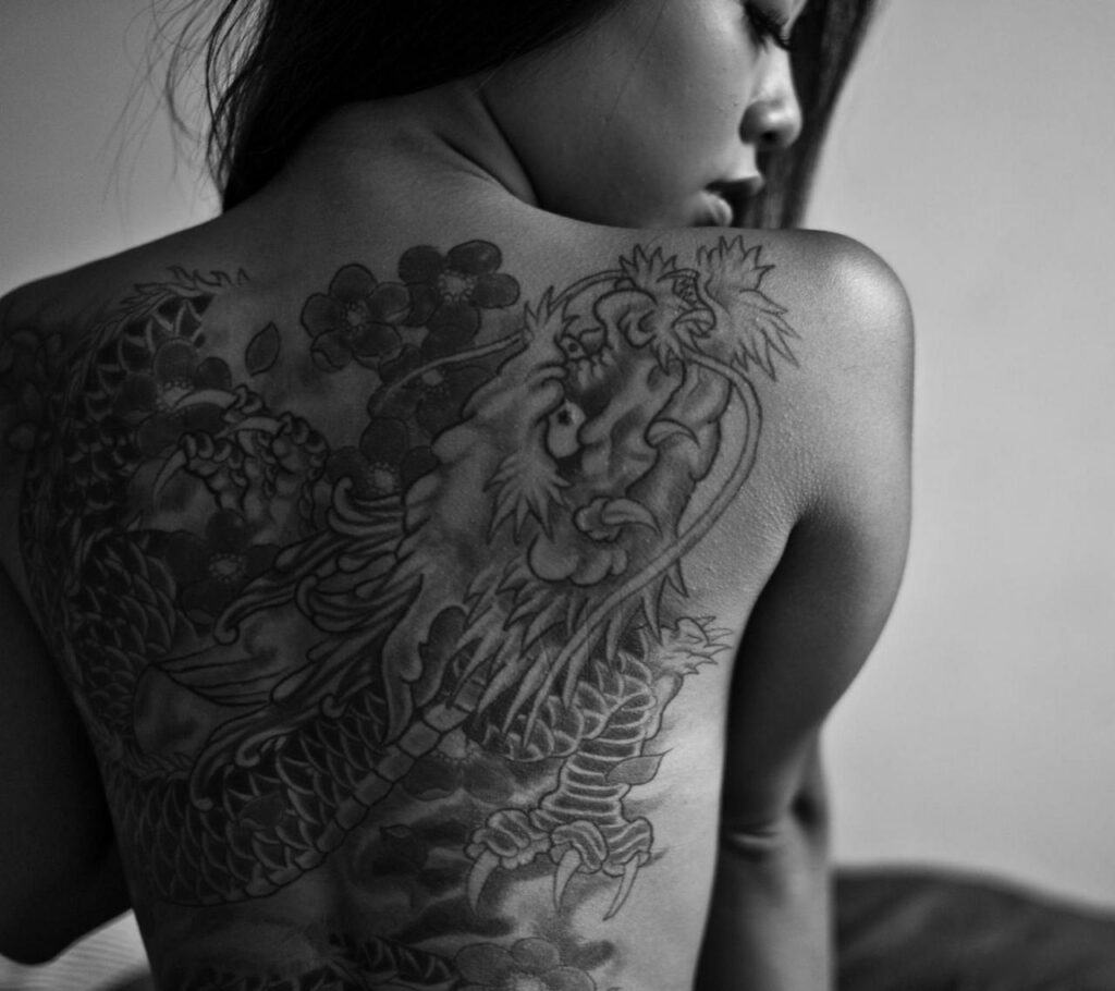 A grayscale portrait of a woman showcasing a detailed back tattoo in monochrome.