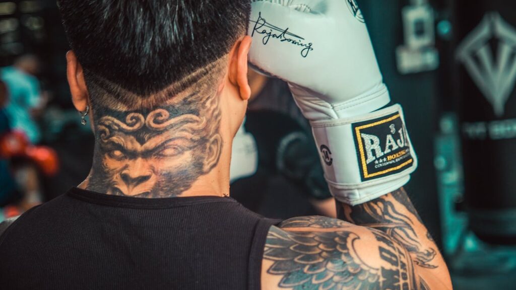 A boxer with intricate tattoos and boxing gloves gets ready for training indoors.